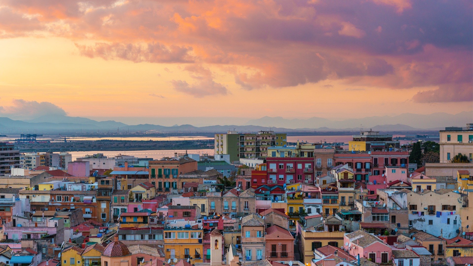 A panoramic view of Cagliari with its colorful buildings under a pink sunset sky. 