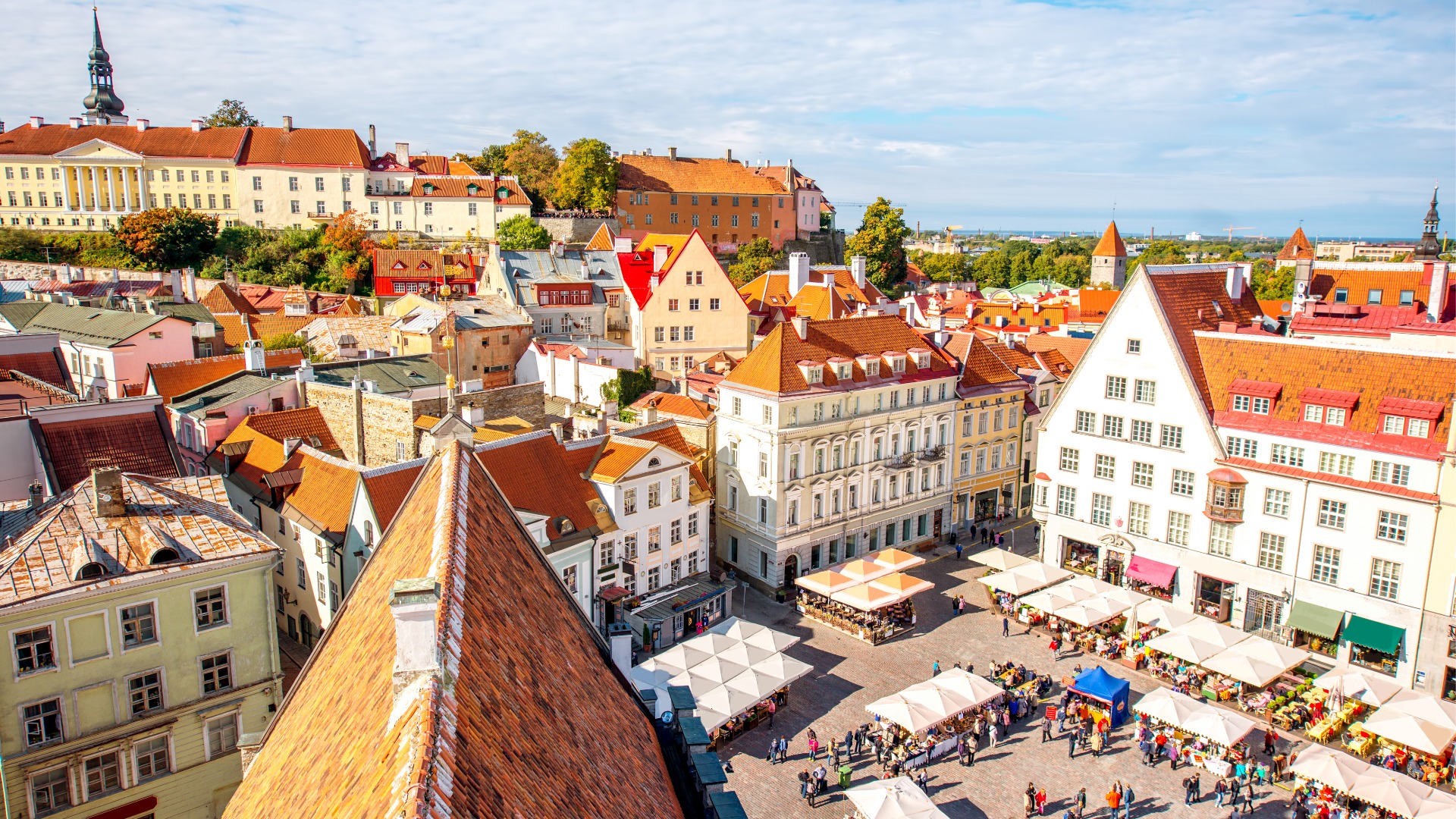 An aerial view of Tallinn's Old Town on a sunny day. The square is filled with people sitting at outdoor cafés. 