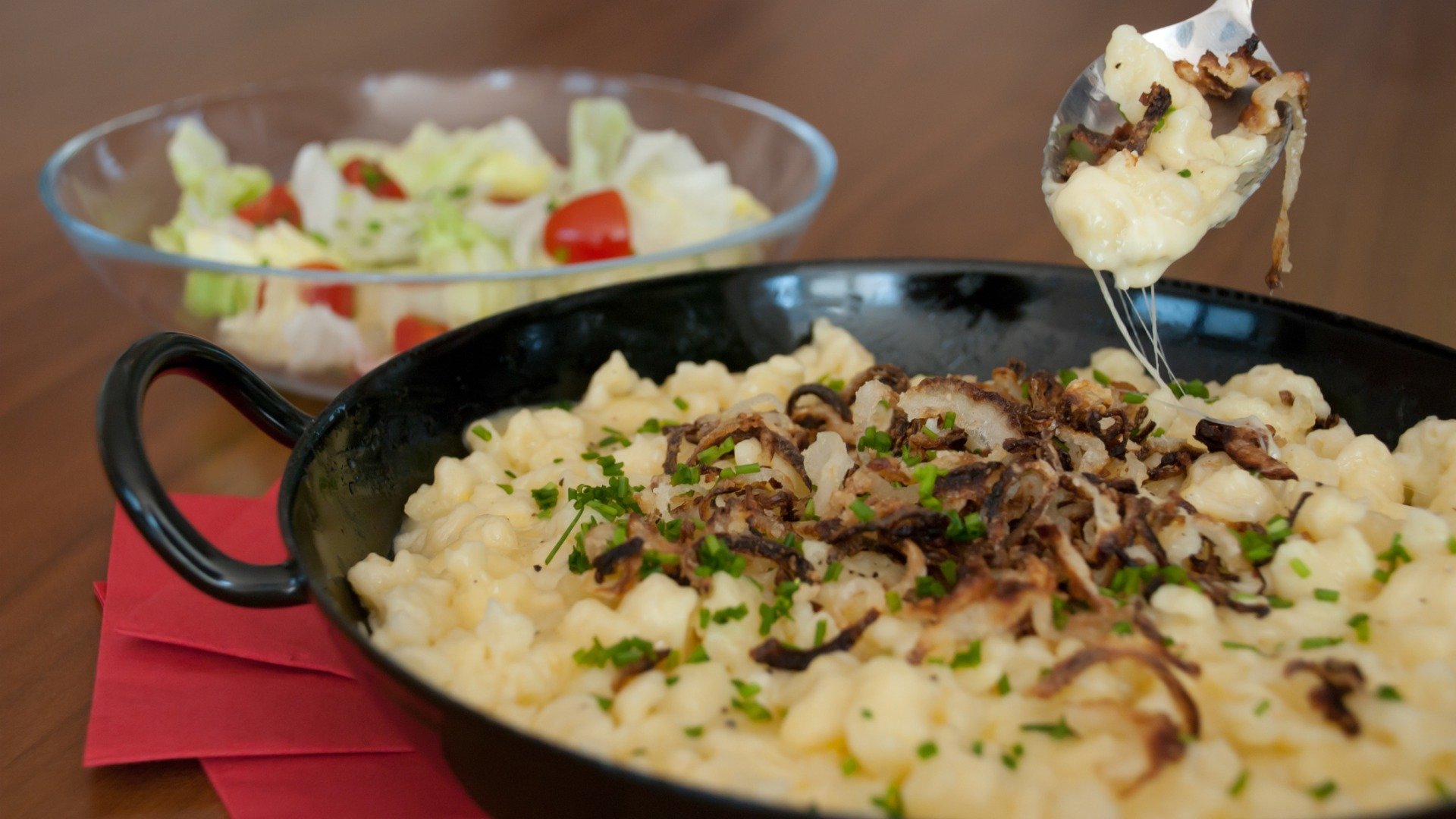 A cast-iron pan filled with pasta and melted cheese. In the background, a small bowl with salad. 