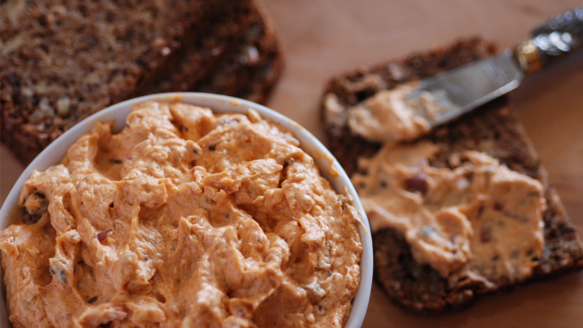 A white bowl filled with an orange dip in the foreground. A knife spreading some of this dip on a slice of rye bread in the background. 