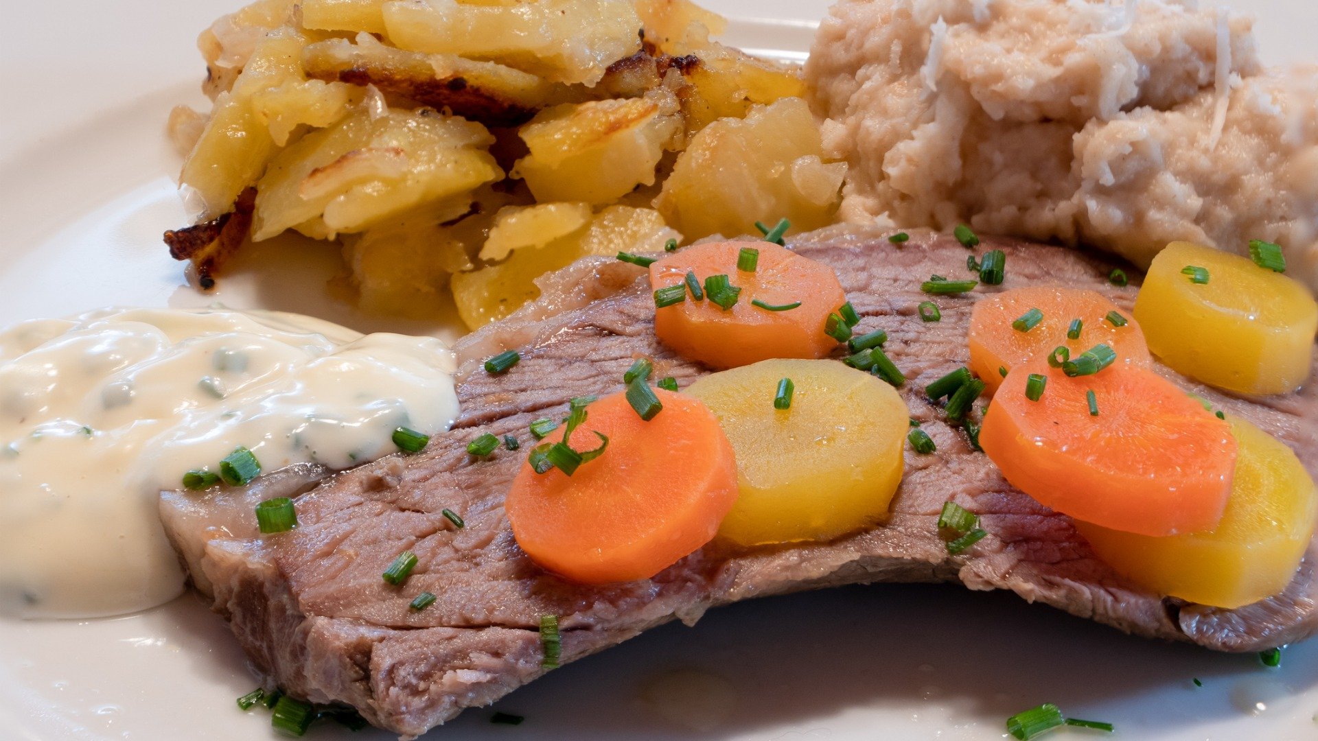 A piece of meat with carrots on top, potatoes and a white sauce as sides. 