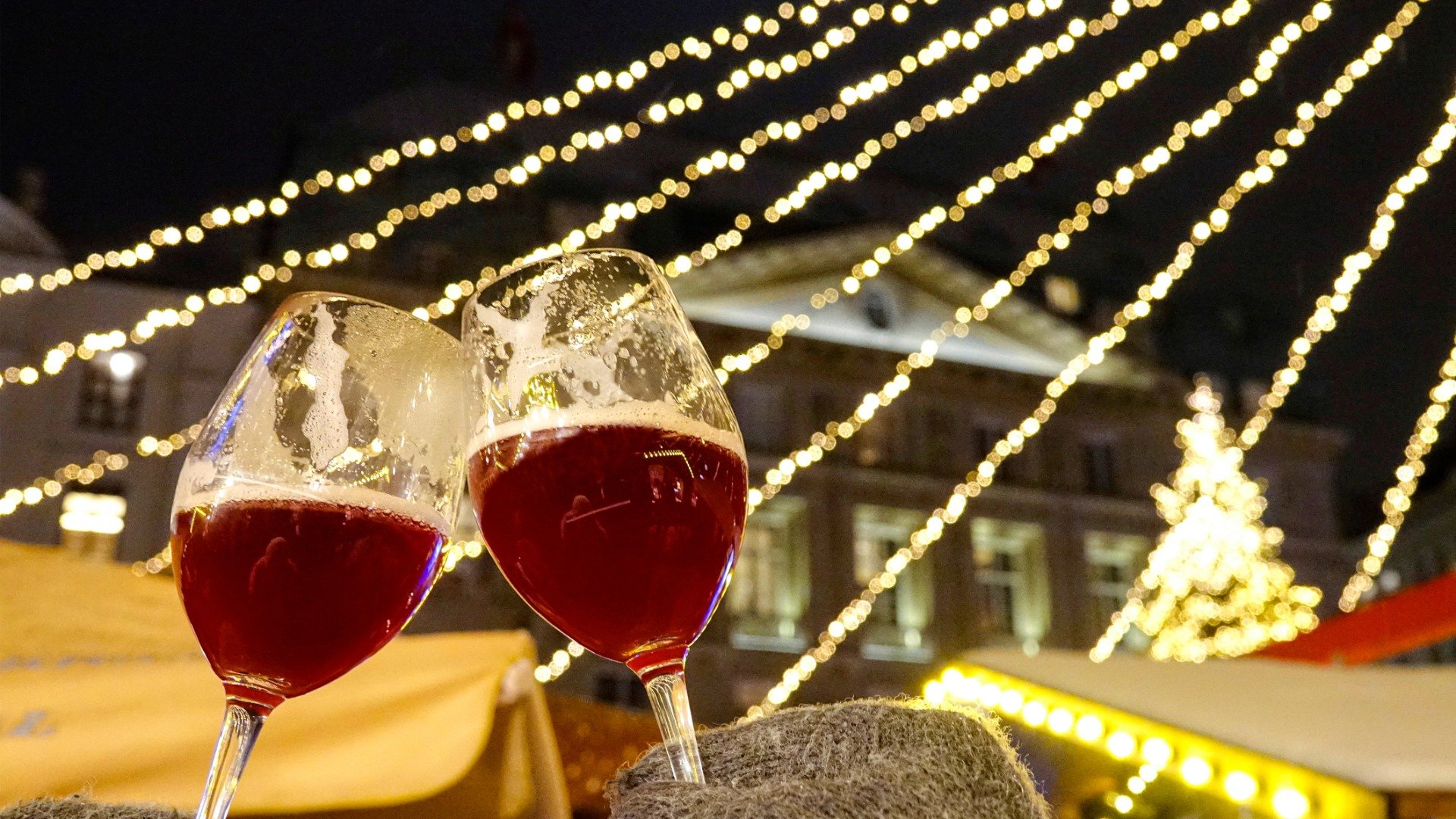 Two tourists wearing grey gloves are toasting with glasses of red mulled wine.