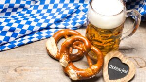 A mug of beer, two pretzels, a wooden heart that has Oktoberfest written on it, and a Bavarian flag in the background.