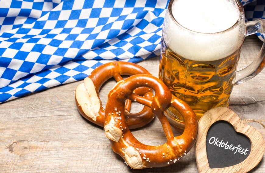 A mug of beer, two pretzels, a wooden heart that has Oktoberfest written on it, and a Bavarian flag in the background.