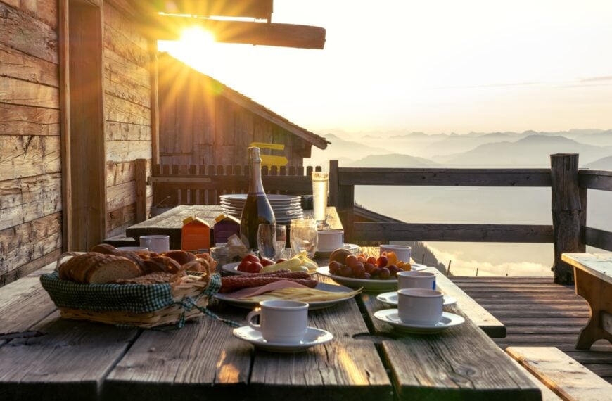 A breakfast table set on a rustic wooden terrace patio of a mountain hut in Tirol at sunrise.