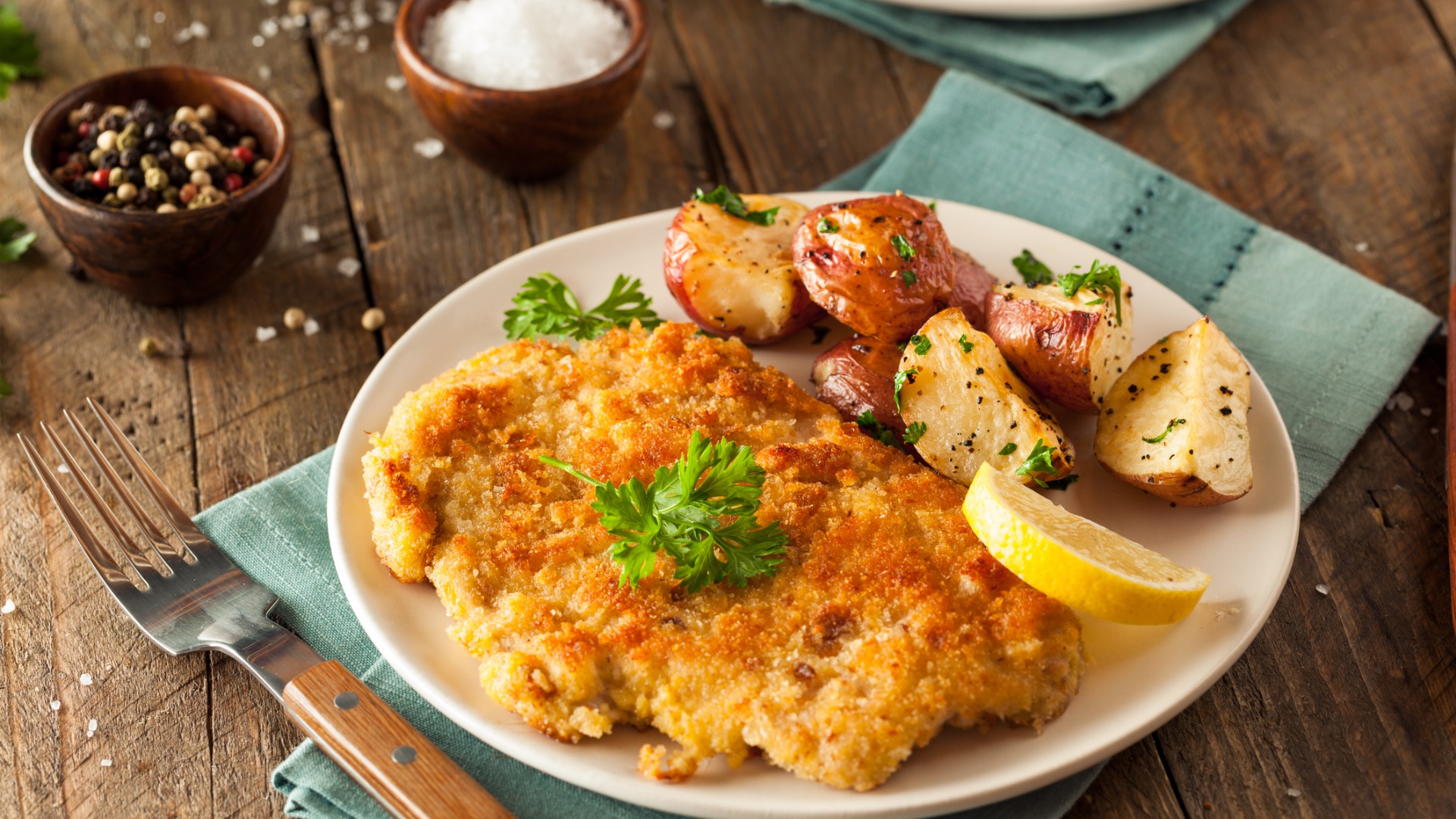 A white dish with a big Schnitzel served with potatoes and a lemon wedge. 