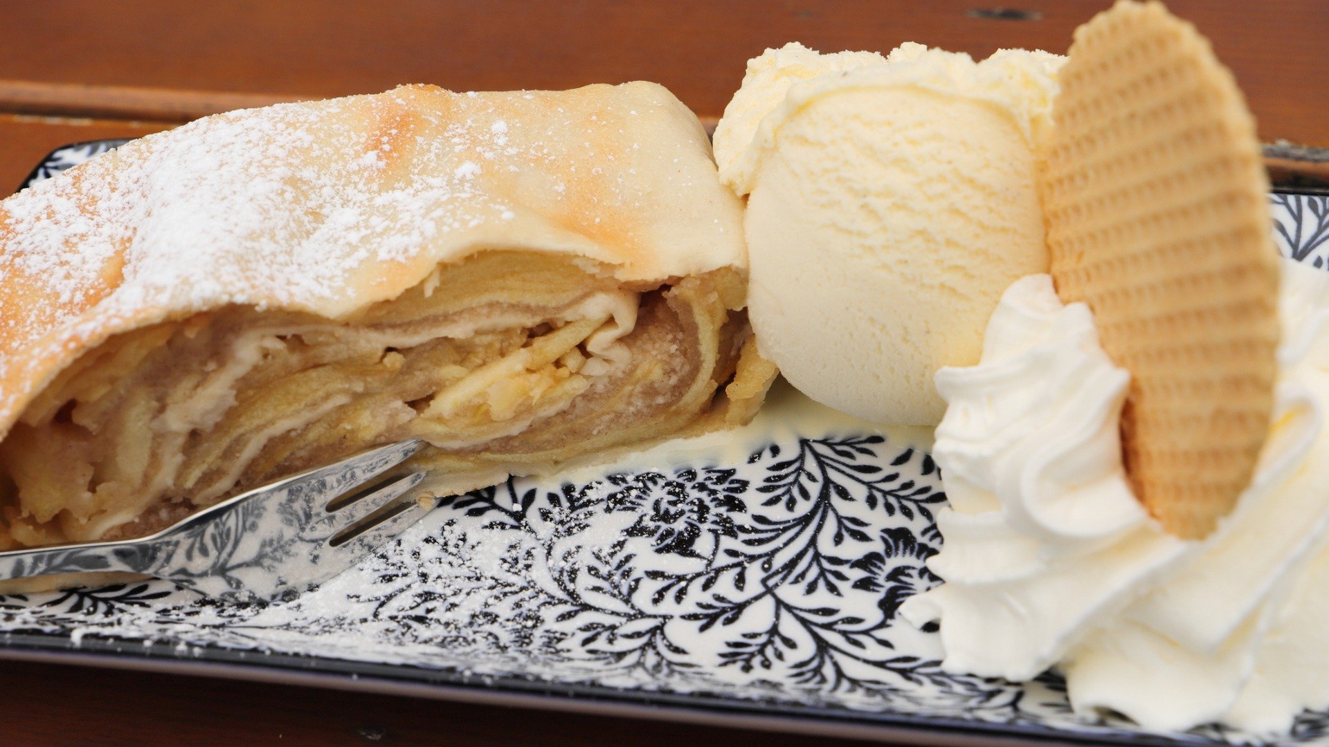 A close up of a piece of apple strudel served with a scoop of vanilla ice cream, a dollop of whipped cream and a thin wafer. 