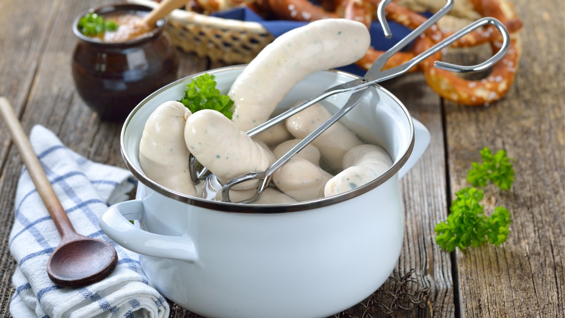 Hot Bavarian white sausages in an enamel cooking pot served on a wooden table with fresh pretzels and sweet mustard.