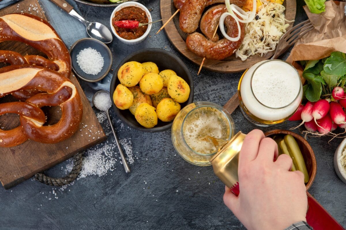 Pretzels, potatoes, sausages and vegetables on a dark surface. A hand is pouring beer from a can into a glass.