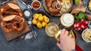 Pretzels, potatoes, sausages and vegetables on a dark surface. A hand is pouring beer from a can into a glass.