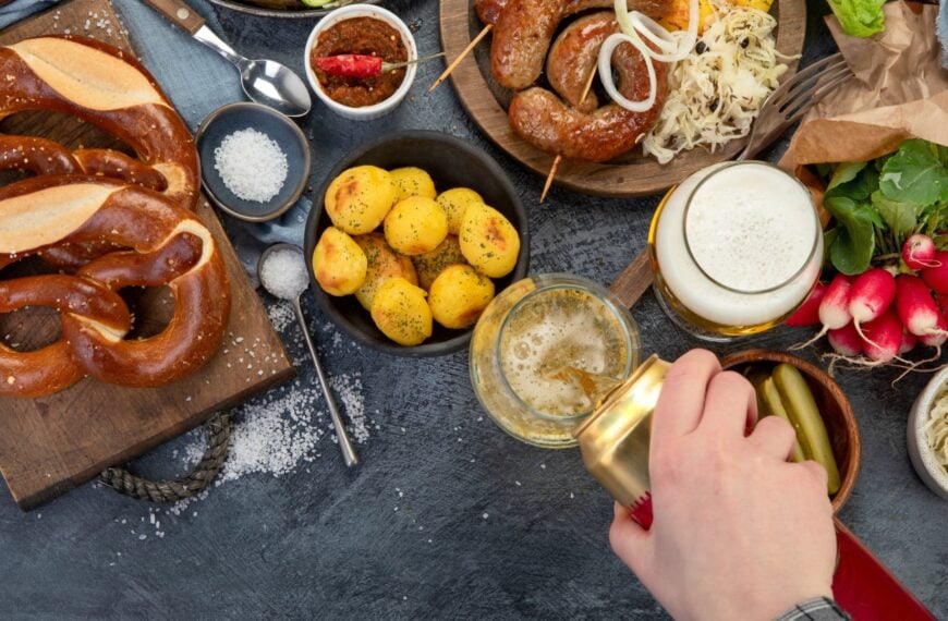 Pretzels, potatoes, sausages and vegetables on a dark surface. A hand is pouring beer from a can into a glass.