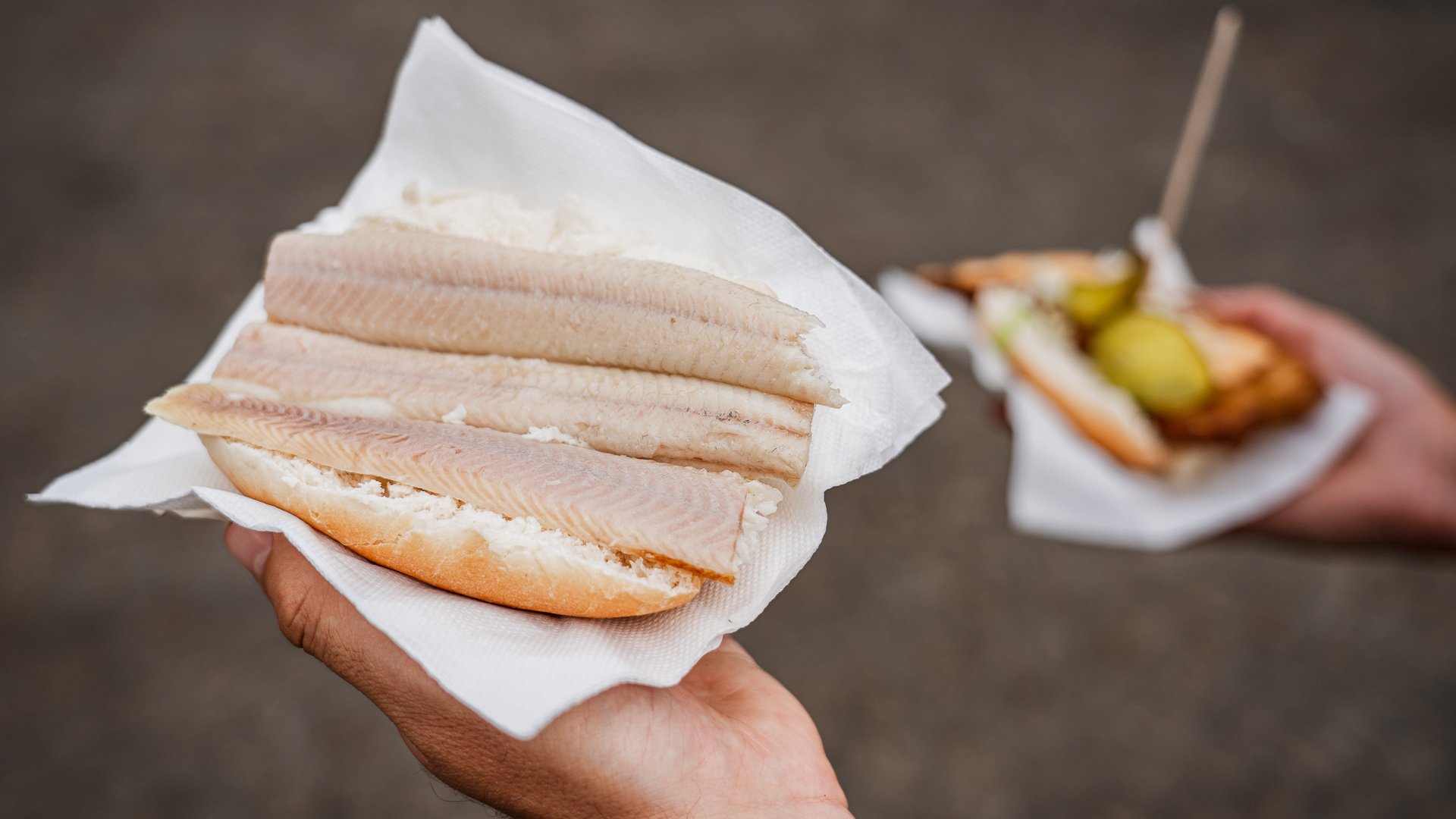 Two hands hold two different fish sandwiches on the streets of Amsterdam.