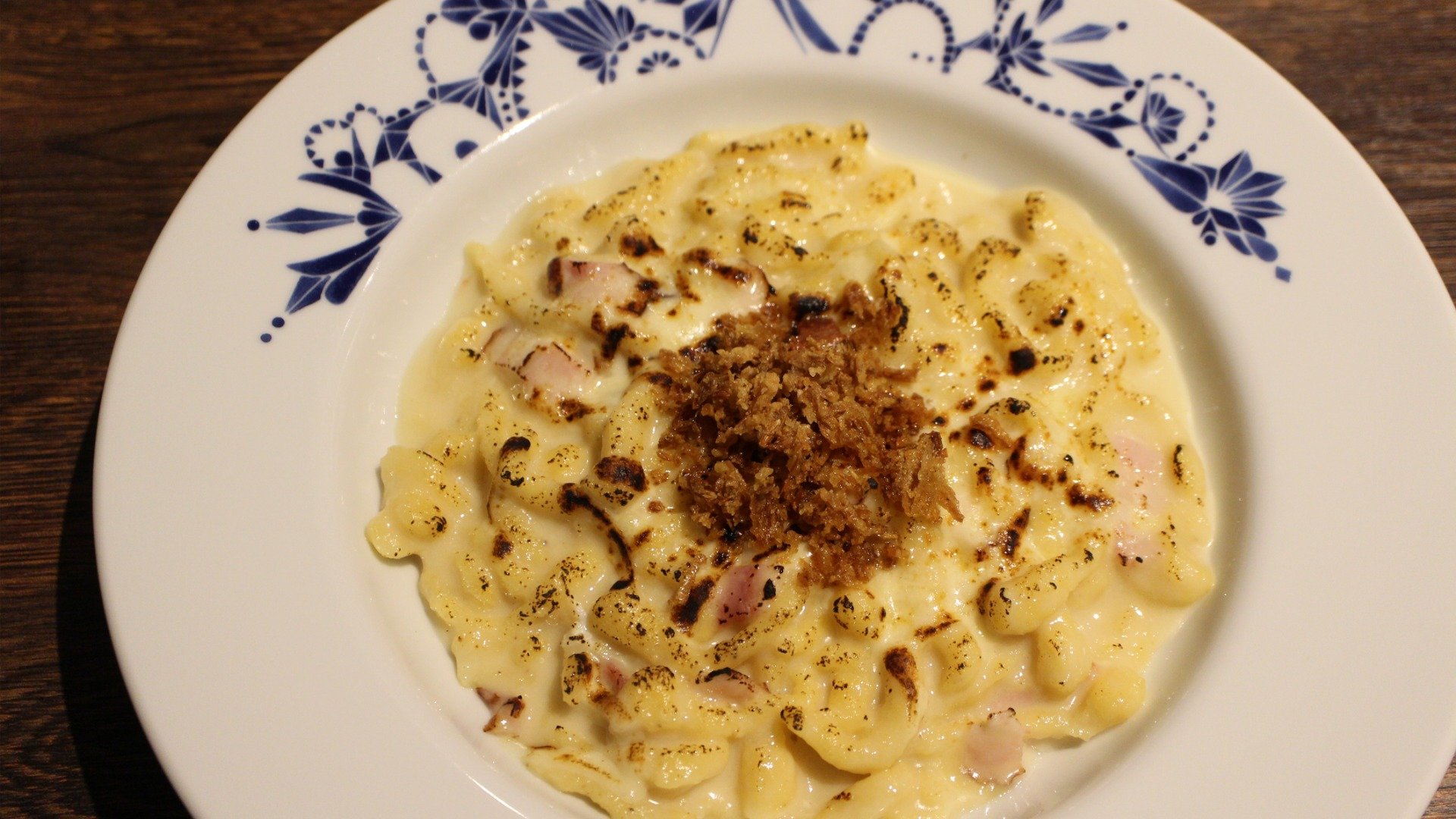 A close up of a white plate with blue painted flowers, filled with Kasespatzle topped with onions. 