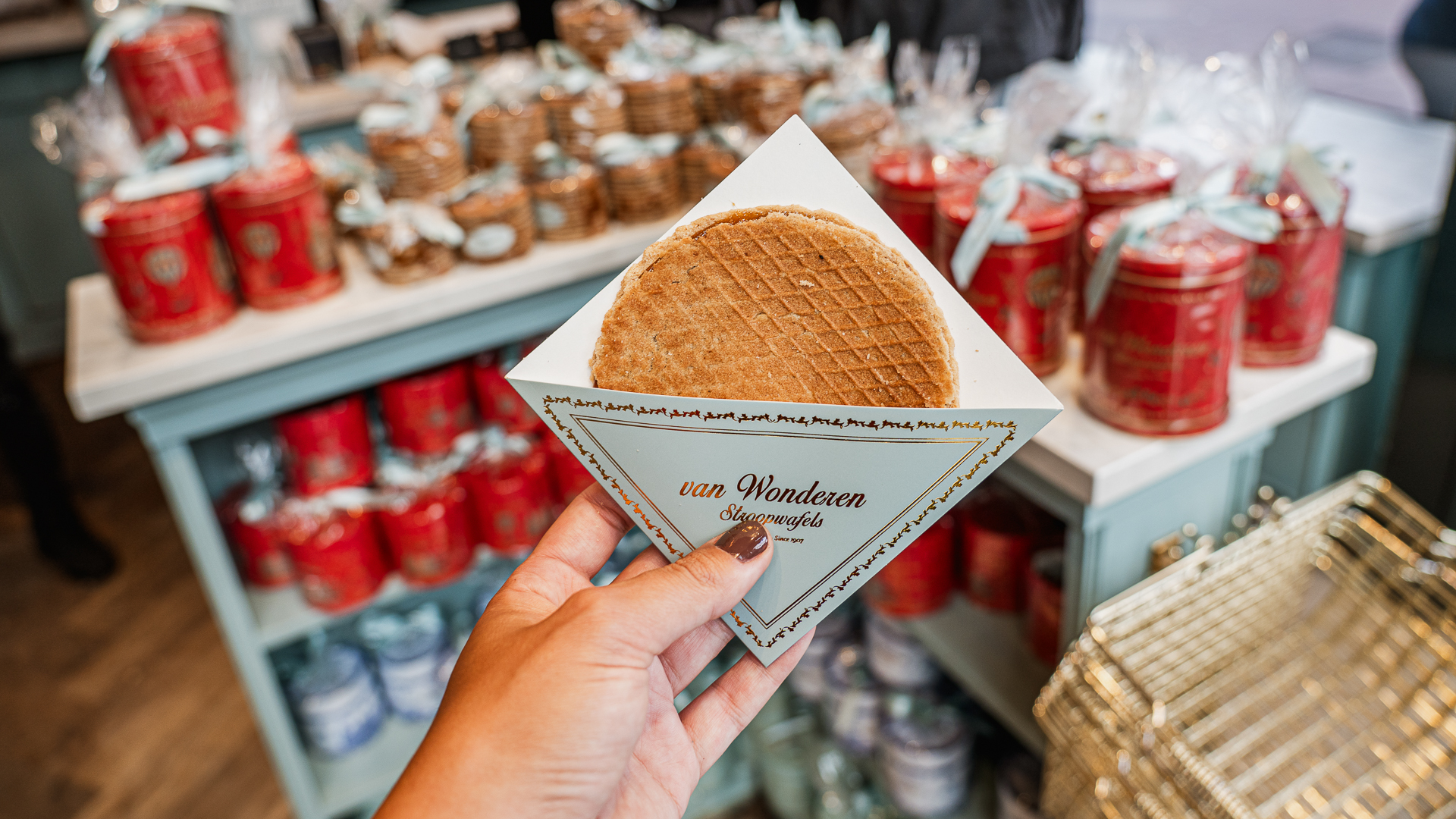 Hand holds a stroopwafel in front of a display of packed stroopwafels.