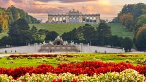 A panoramic view of the Gloriette overlooking the Schonbrunn Gardens under a glorious sunset sky. There are flowers of vivid colors in the foreground.