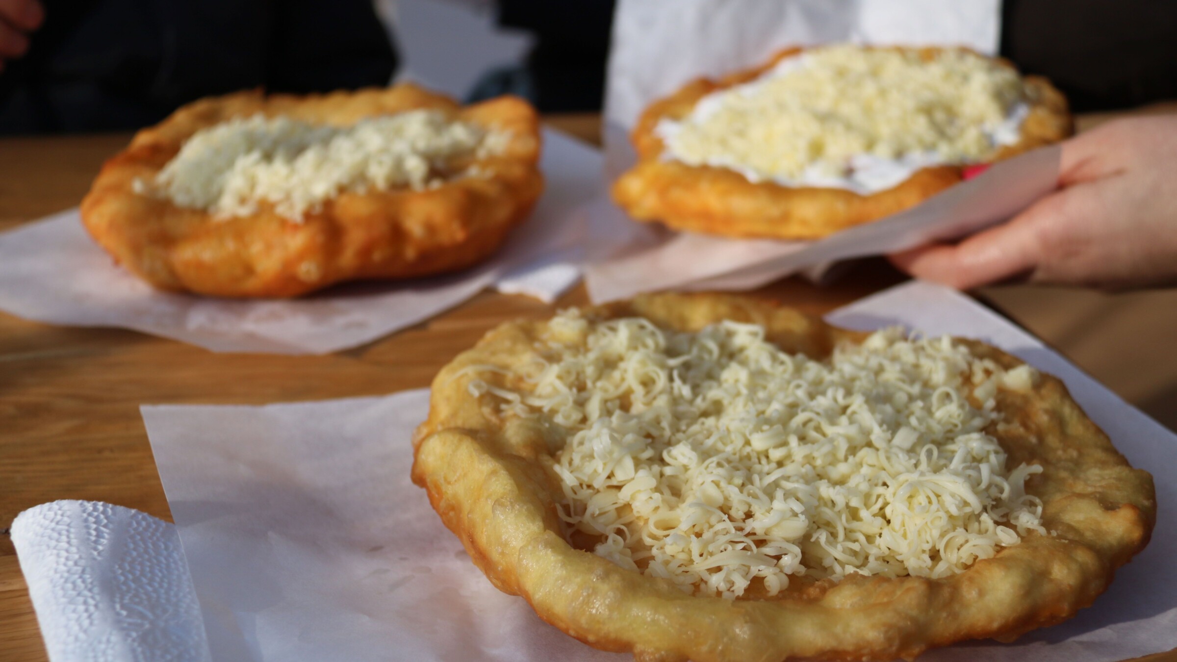 Three langos topped with grated cheese and served on sheets of paper.