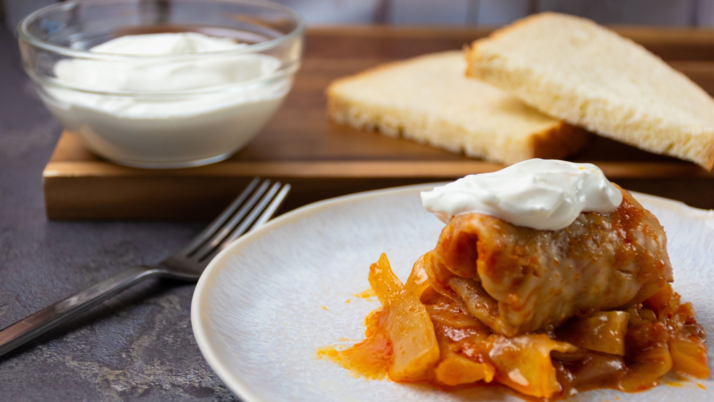 A Hungarian stuffed cabbage roll with a small bowl of sour cream and two slices of bread in the background.