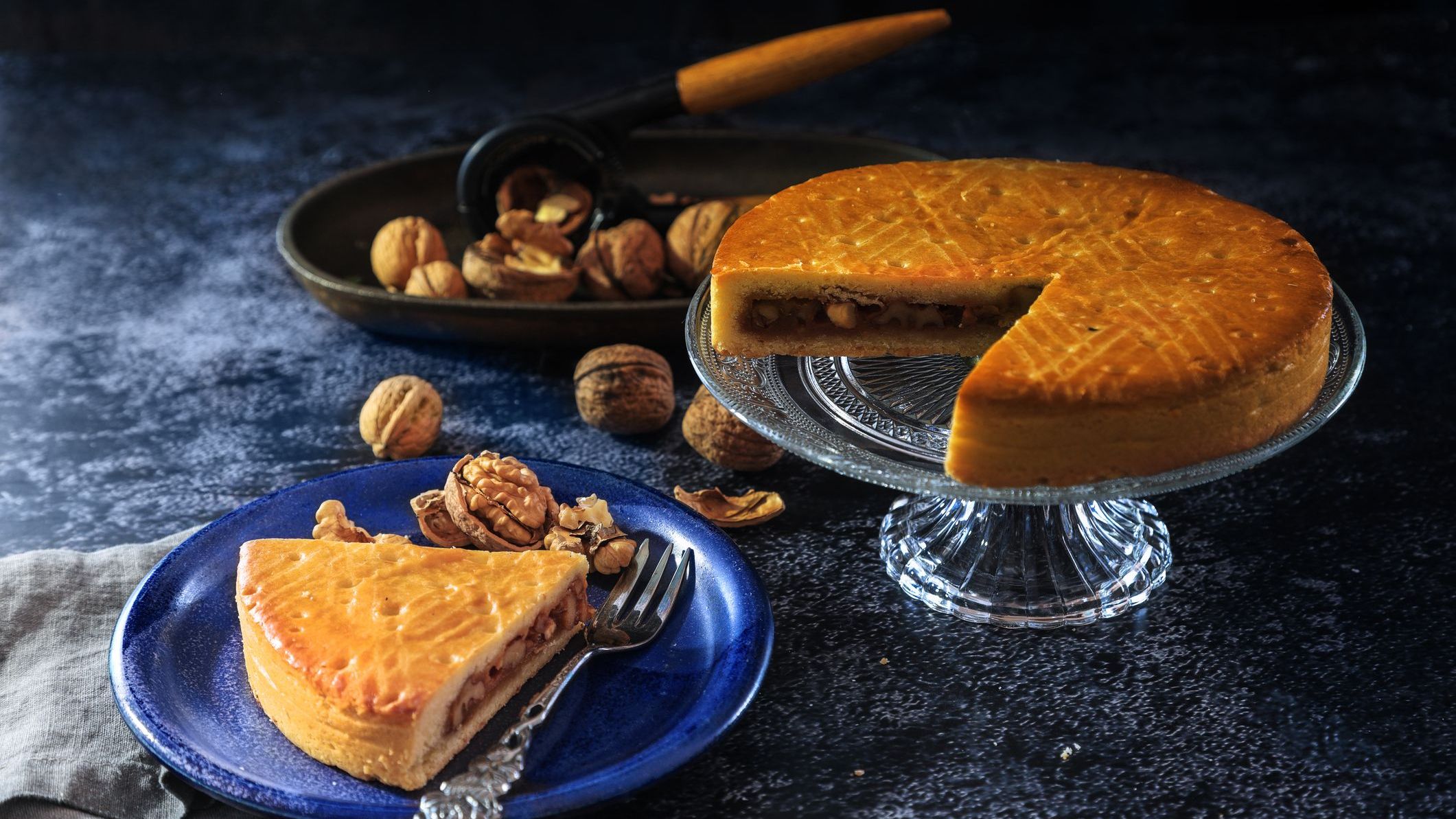 An Engadine Nusstorte cake on a glass cake stand and a piece of Engadine Nusstorte served on a blue plate with walnuts in the background.