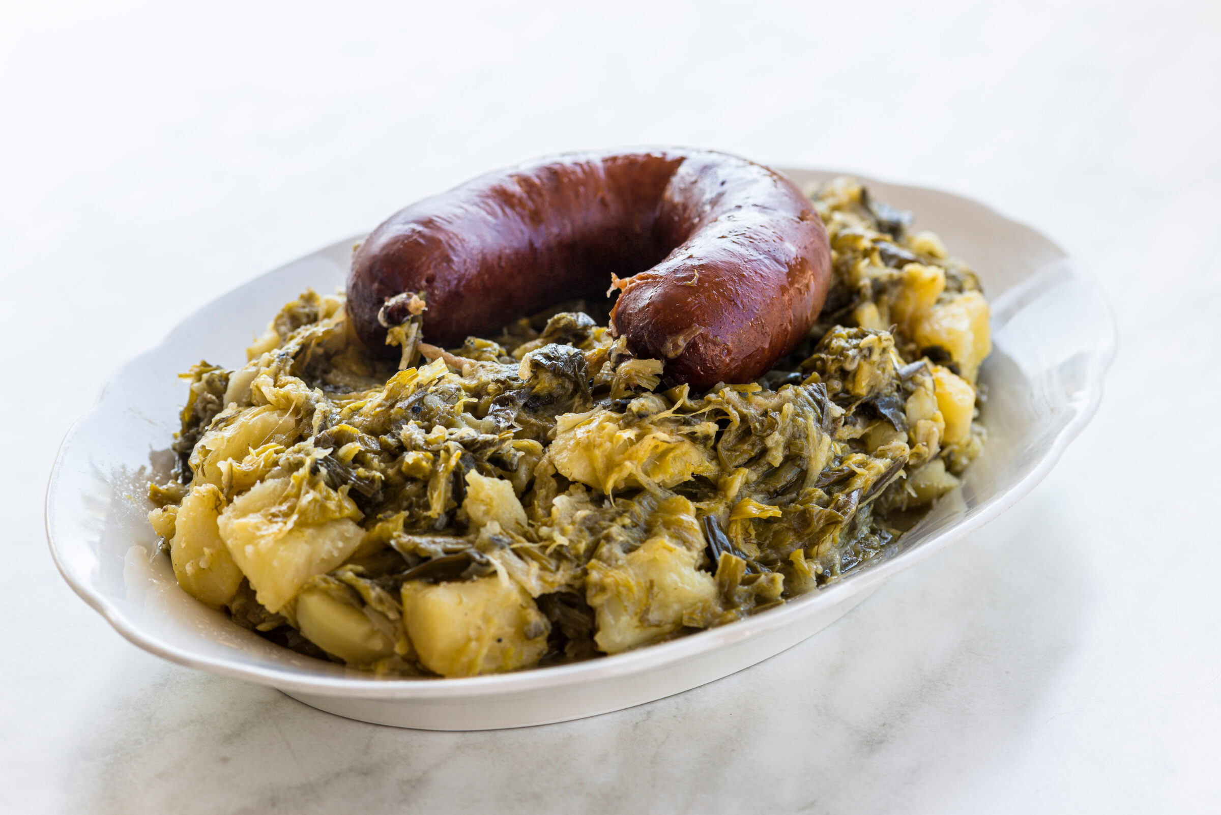 A white plate with potatoes and leek, topped with a sausage.