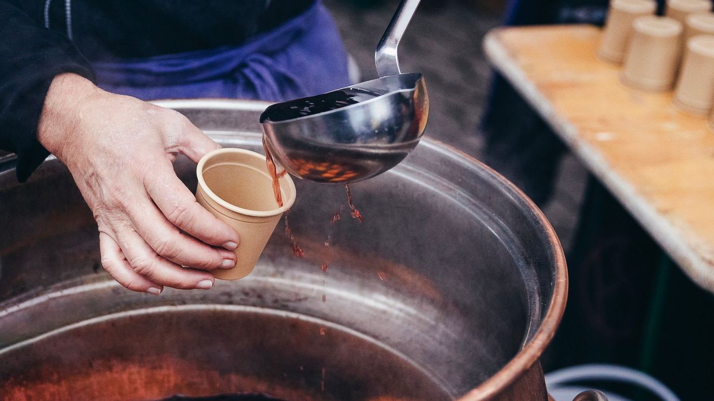 Pouring hot mulled wine into a paper cup using a huge kitchen spoon.
