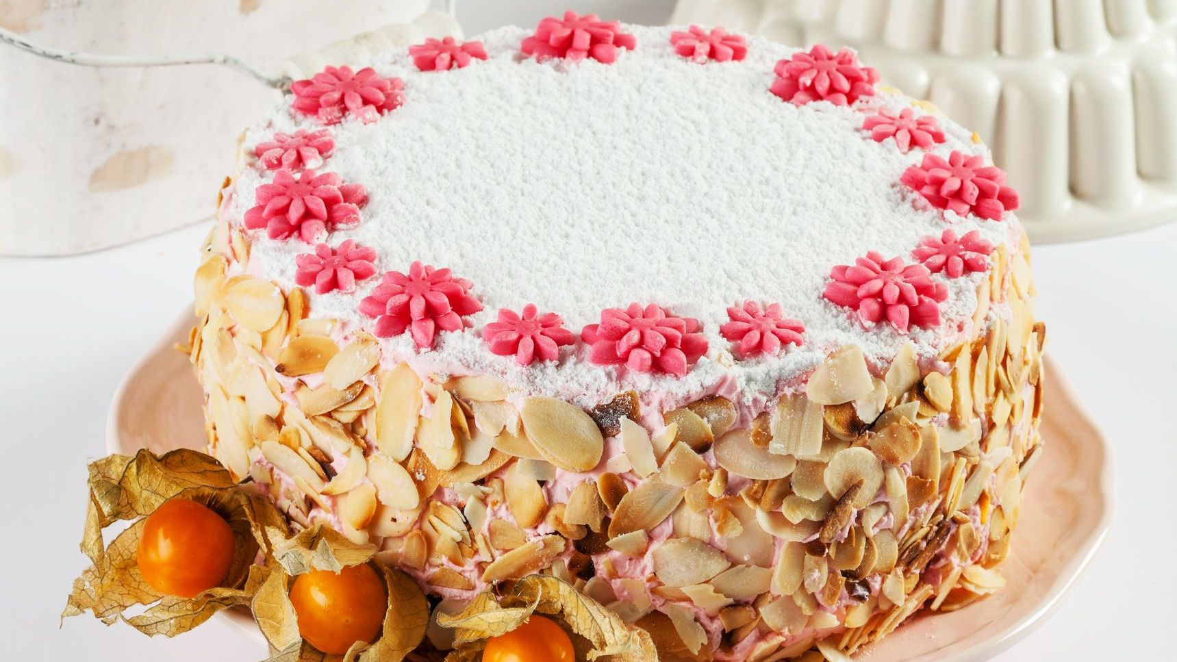 A white cake topped with icing sugar and pink candy flowers and covered with almond flakes on its sides.