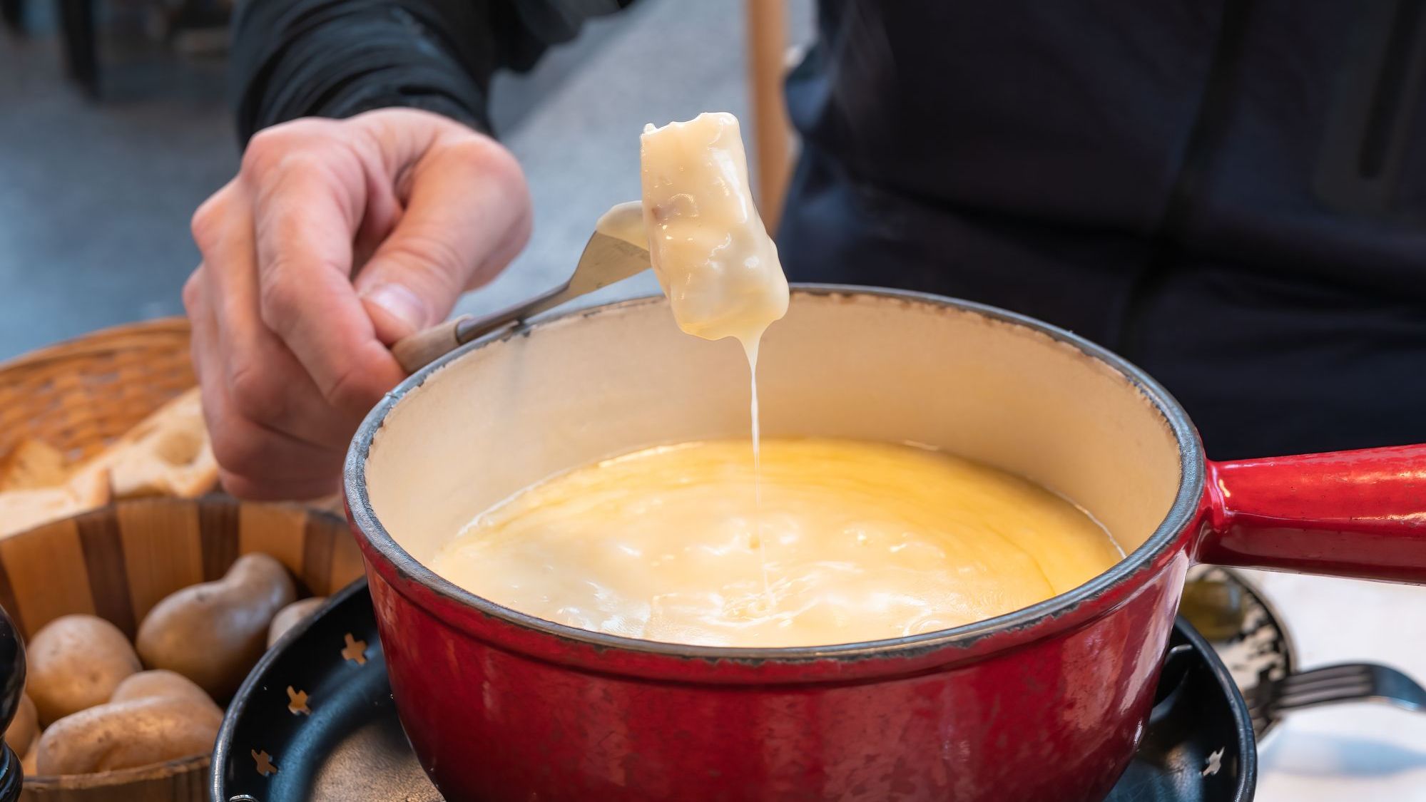 A man's hand holds a fork with a piece of bread that has been dipped in a cheese fondue pot.