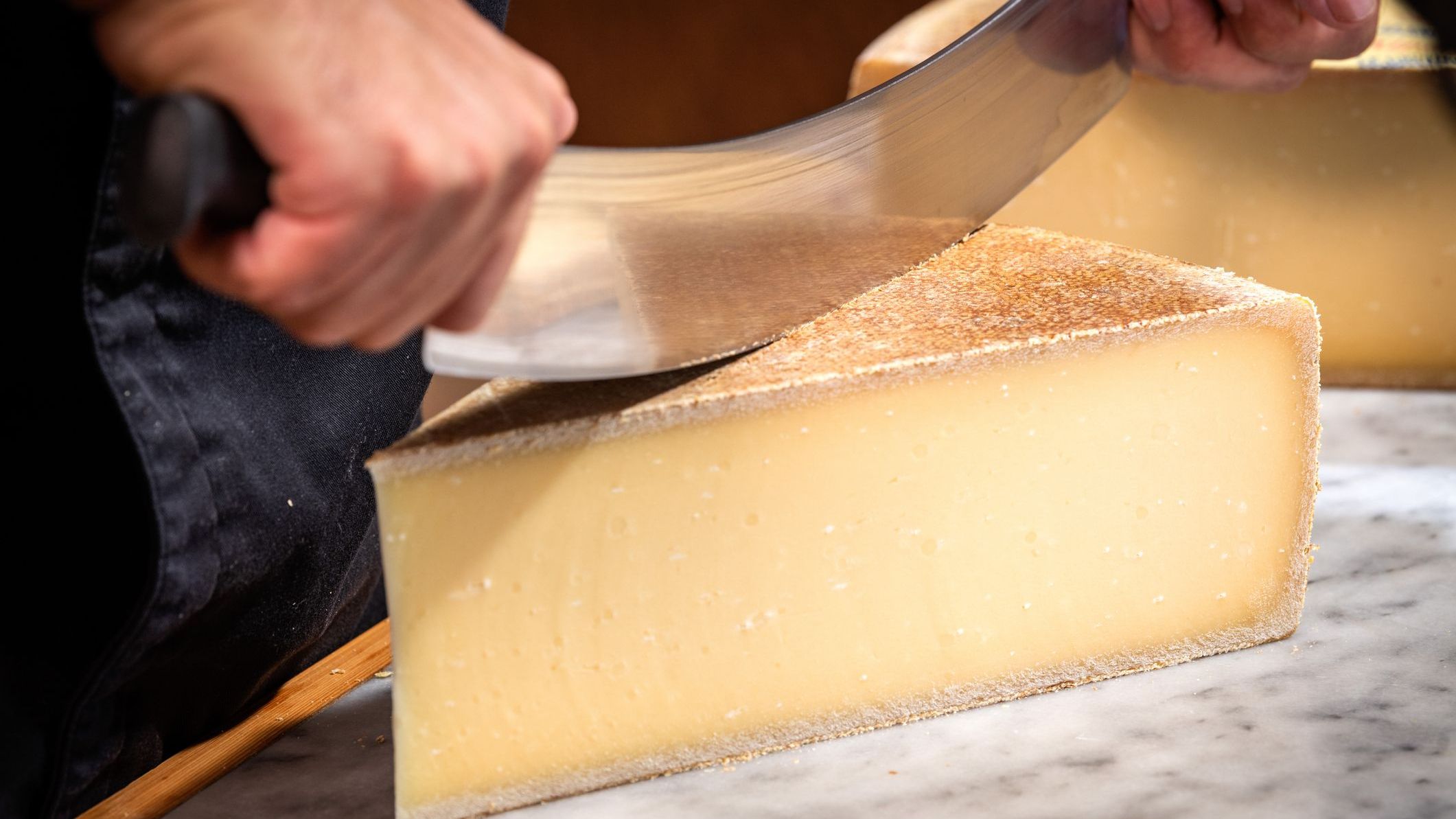 A man is cutting a portion of Gruyere with a large knife on a marble background in a cellar.