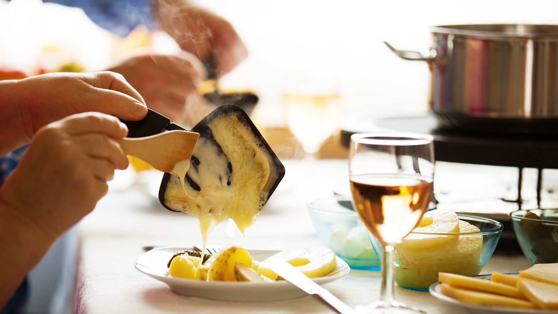 Two hands are scraping melted Raclette cheese on boiled potatoes in a white plate.