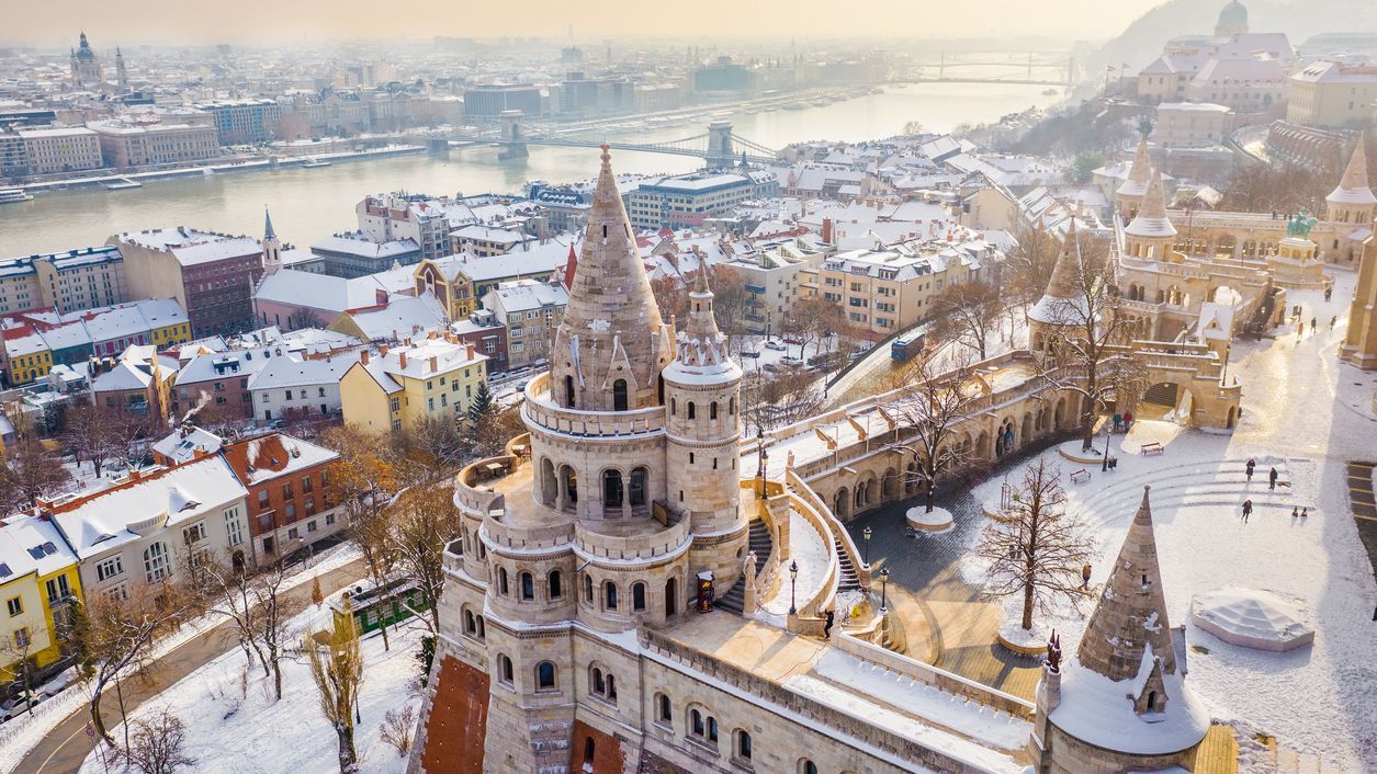 Aerial view of the snowy Fisherman's Bastion with the Szechenyi Chain Bridge and St. Stephen's Basilica in the background on a snowy winter morning.