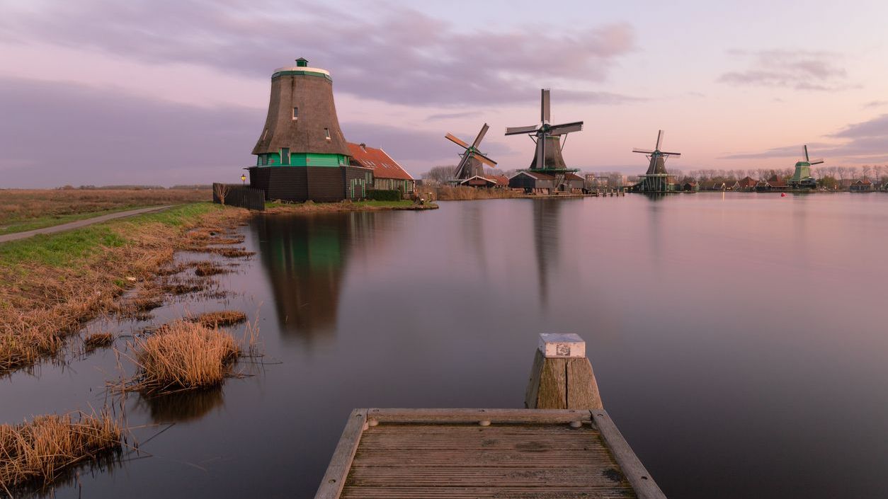 A dreamy landscape of wooden windmills by the water under a lilac sky.