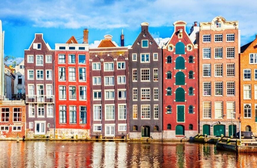 A row of typical Amsterdam buildings of various colors, lining a canal.