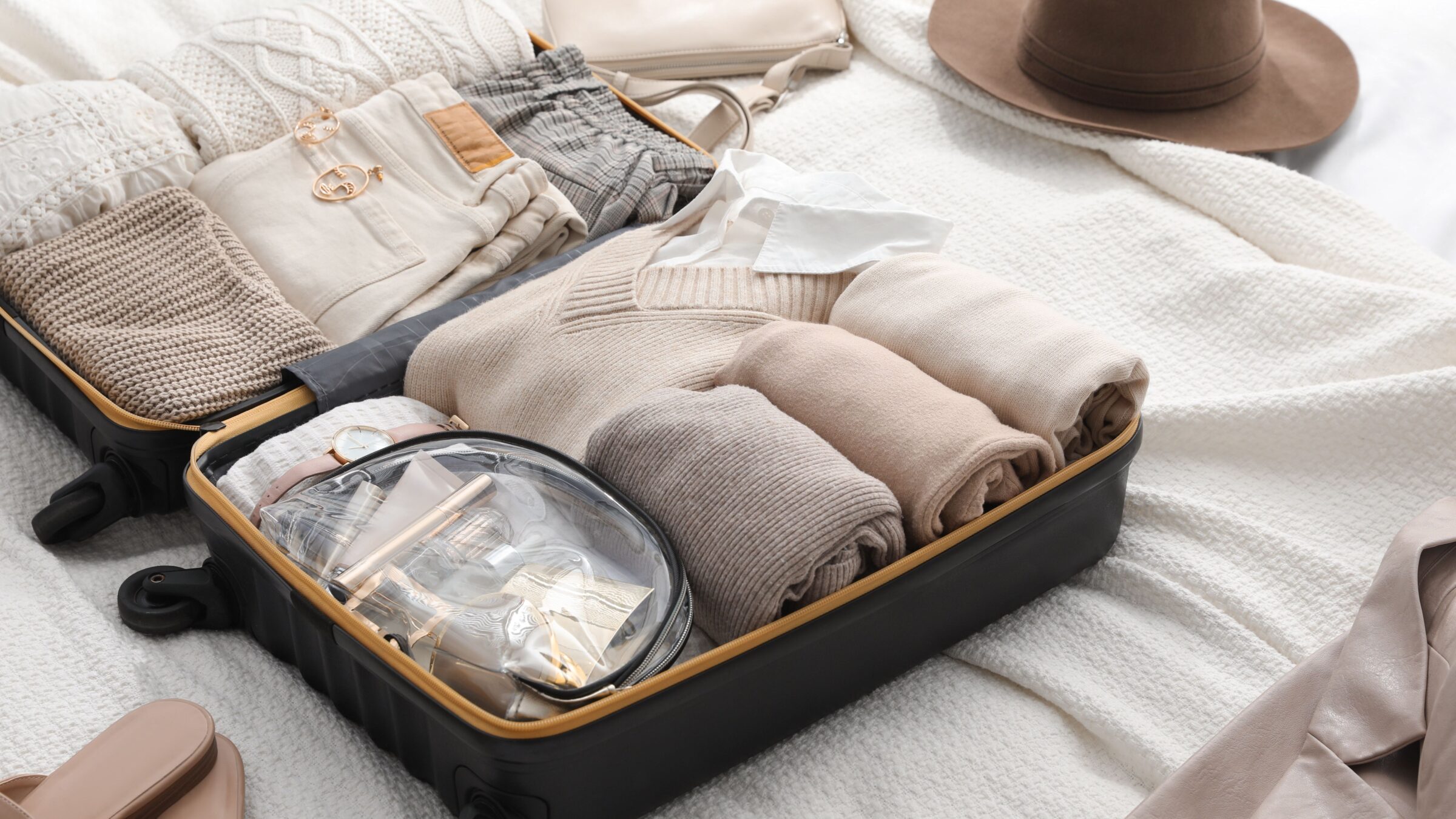 An open suitcase with folded clothes, shoes and accessories on a bed.
