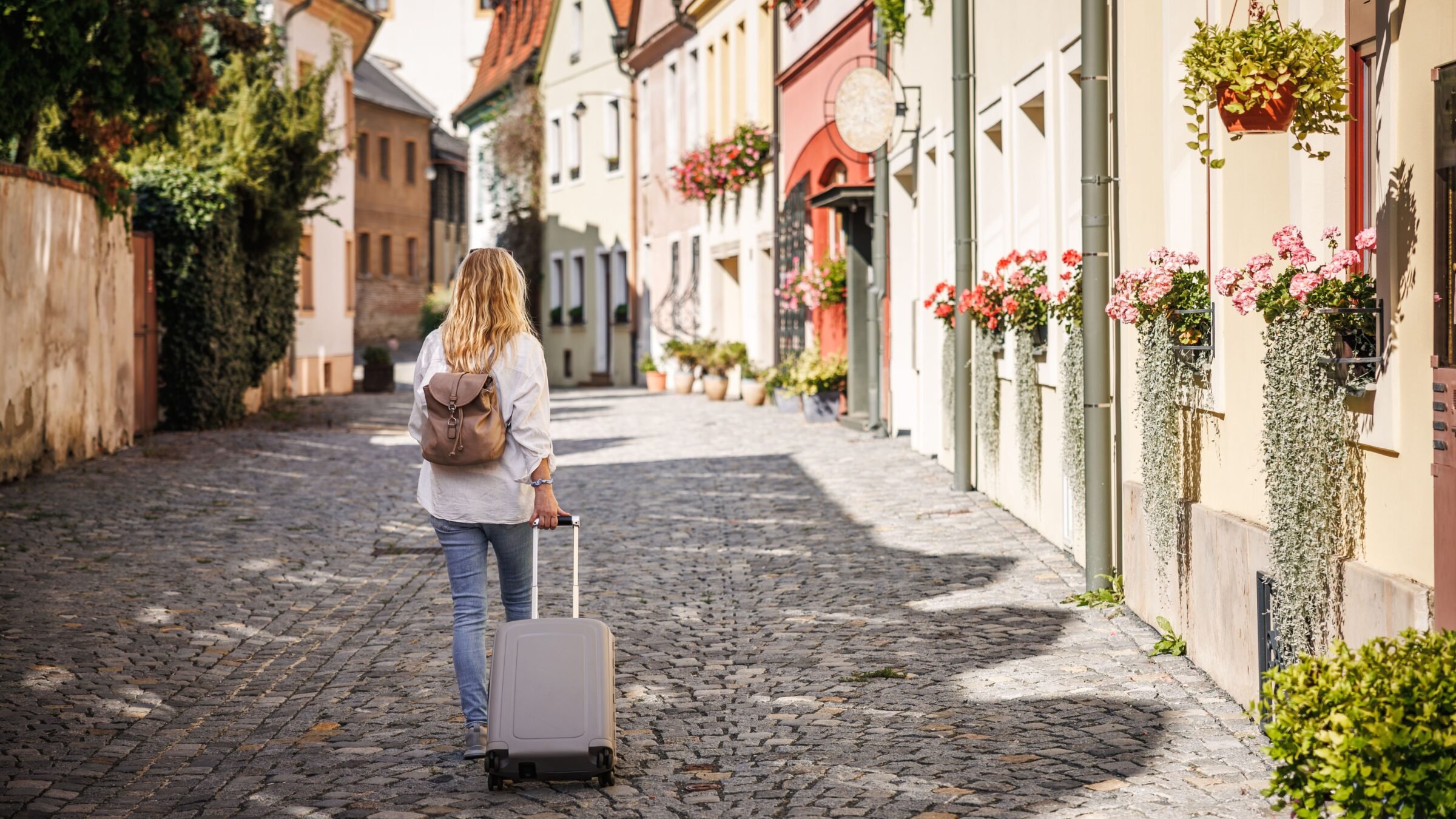 A woman with a suitcase and a small backpack walking along a cobbled street lined with beautiful buildings.