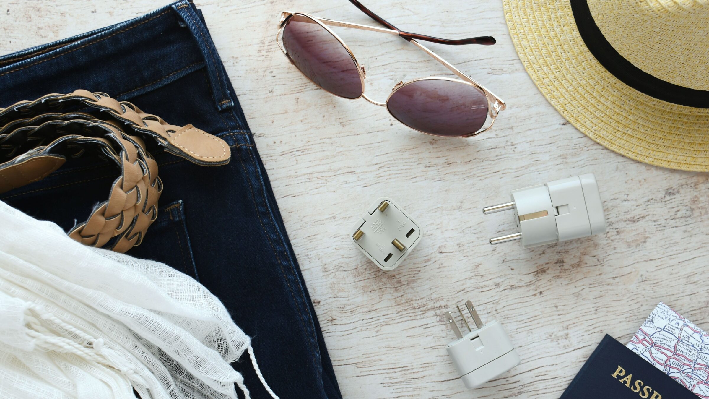 Travel essentials flat lay, including a power converter, a voltage travel adapter, sunglasses, a straw hat, a belt, a passport with a folded map inside, a pair of jeans, and a scarf.