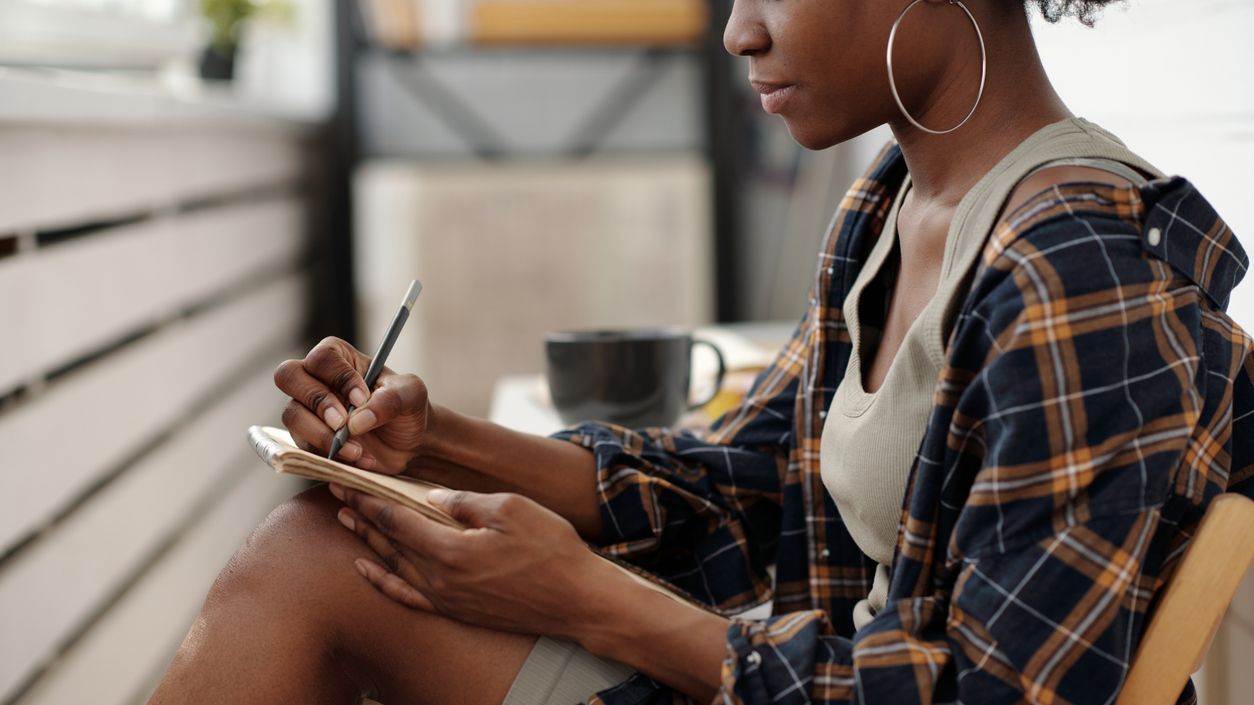 A woman is writing notes while sitting indoors. She's holding a pen in one hand and a notebook in the other.