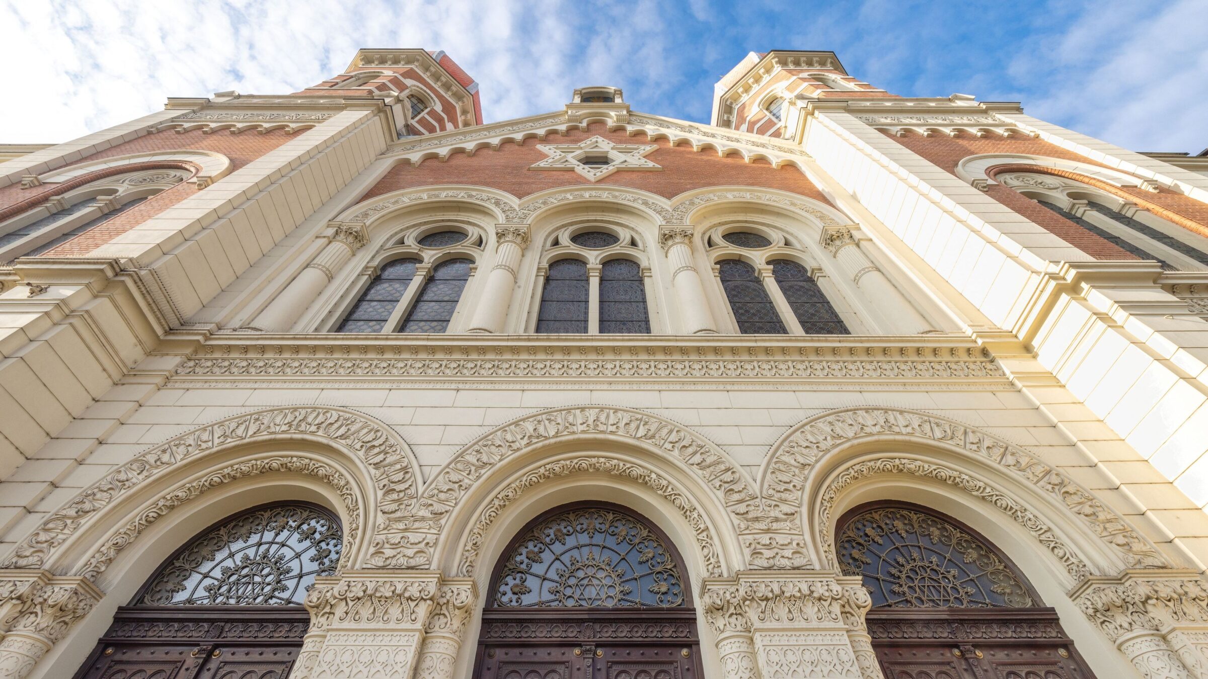 An architectural marvel: intricate detailing adorns this grand building's facade, featuring arched doorways, ornate stonework, and elegant windows. The warm tones of the brick and the light-colored stone create a striking visual contrast against a bright blue sky.