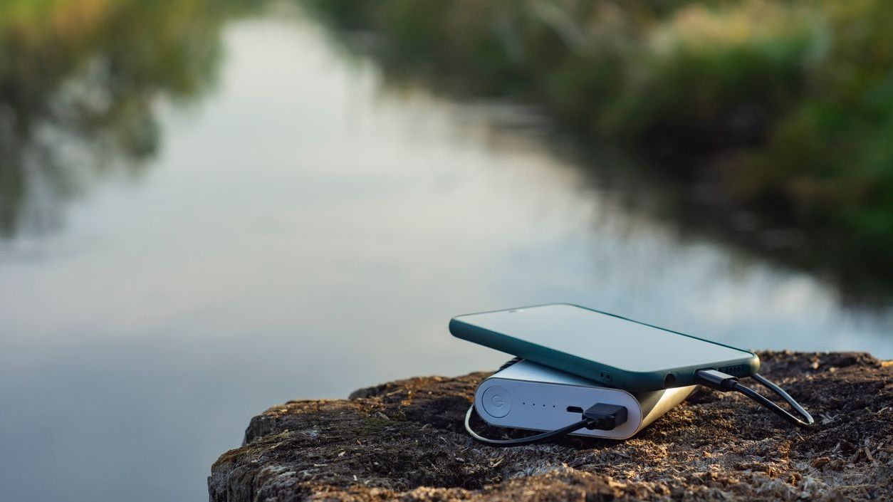 A smartphone is charged using a portable charger outdoors with a river in the background.