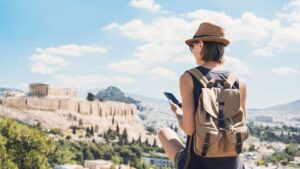 A young woman with her back turned to the camera is looking at her phone. The Acropolis of Athens and Lycabettus Hill in the background.