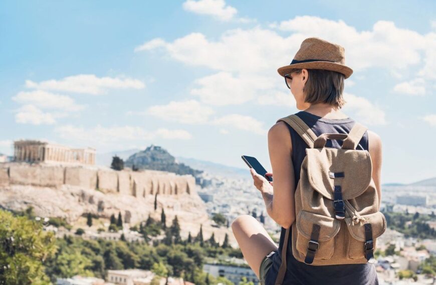 A young woman with her back turned to the camera is looking at her phone. The Acropolis of Athens and Lycabettus Hill in the background.