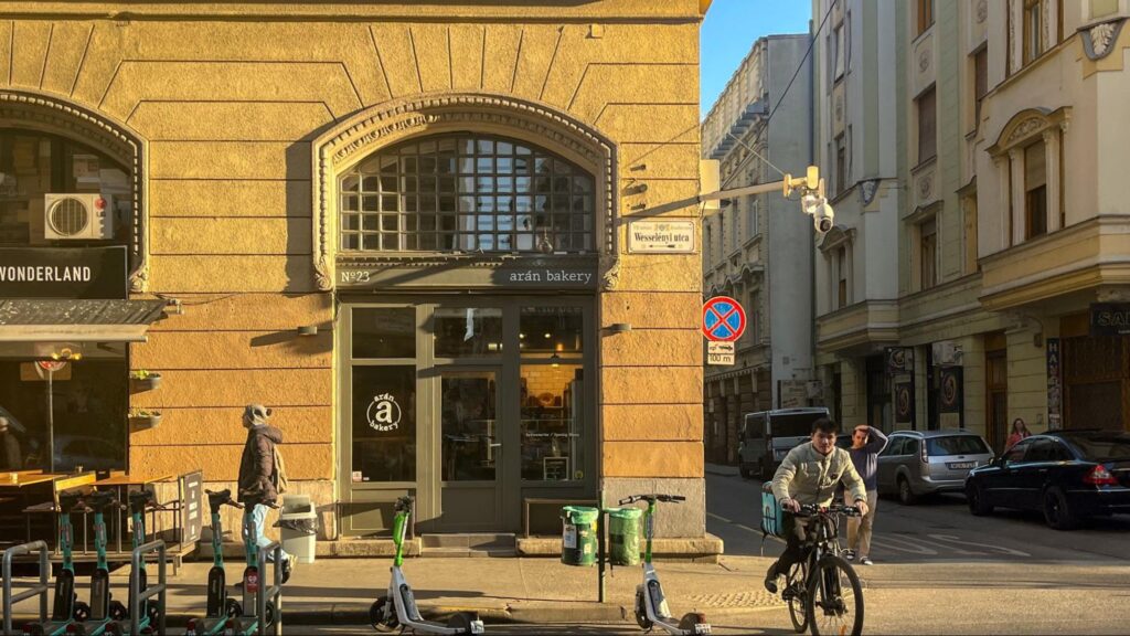 Street view of Arán Bakery located on Wesselényi utca in Budapest, with a warm-toned historic facade, arched windows, and scooters parked out front. Pedestrians and a cyclist pass by in the late afternoon sunlight
