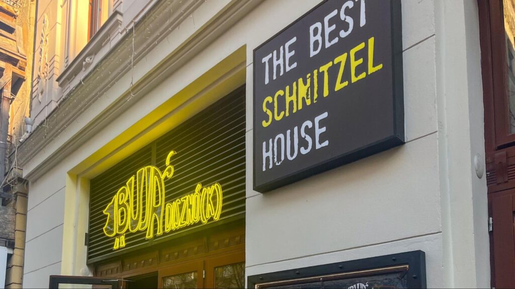 Street view of the entrance to BUNSZNÓ(K), a restaurant with a bright yellow neon sign and a bold black-and-white sign reading 'The Best Schnitzel House' mounted on a white building facade