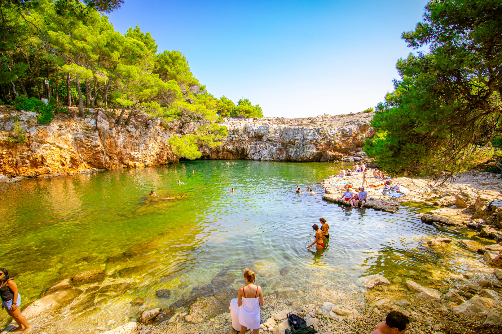 People swimming and relaxing around the vibrant green waters of the Dead Sea (Mrtvo More) lagoon on Lokrum Island, surrounded by rocky cliffs and pine trees.