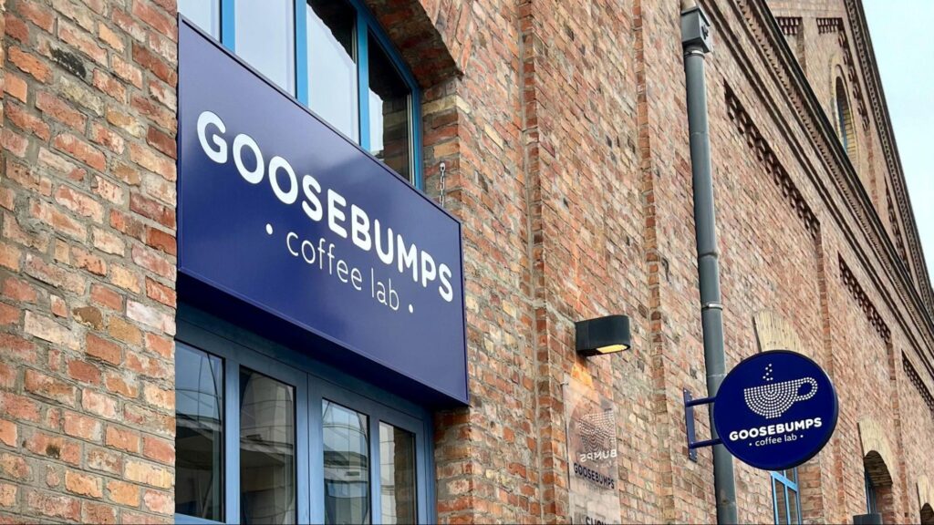 Exterior of Goosebumps Coffee Lab, a specialty coffee shop with a modern blue sign on a historic brick building. The logo features a minimalist coffee cup and steam design."