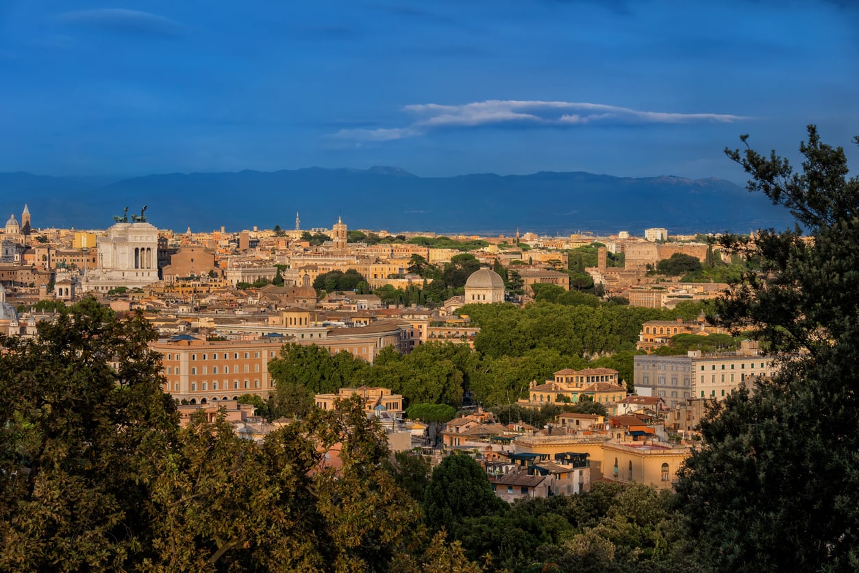 Panoramic view of Rome at sunset with the Altare della Patria, domes of historic churches, and tree-lined rooftops, set against the backdrop of distant mountains