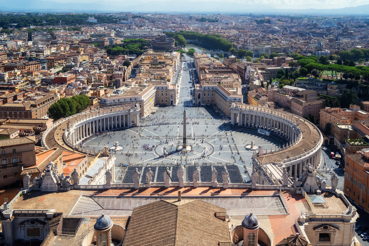Aerial view of St. Peter’s Square in Vatican City, featuring the central obelisk, colonnades, and the surrounding buildings of Rome, with Castel Sant’Angelo and the Tiber River in the background.