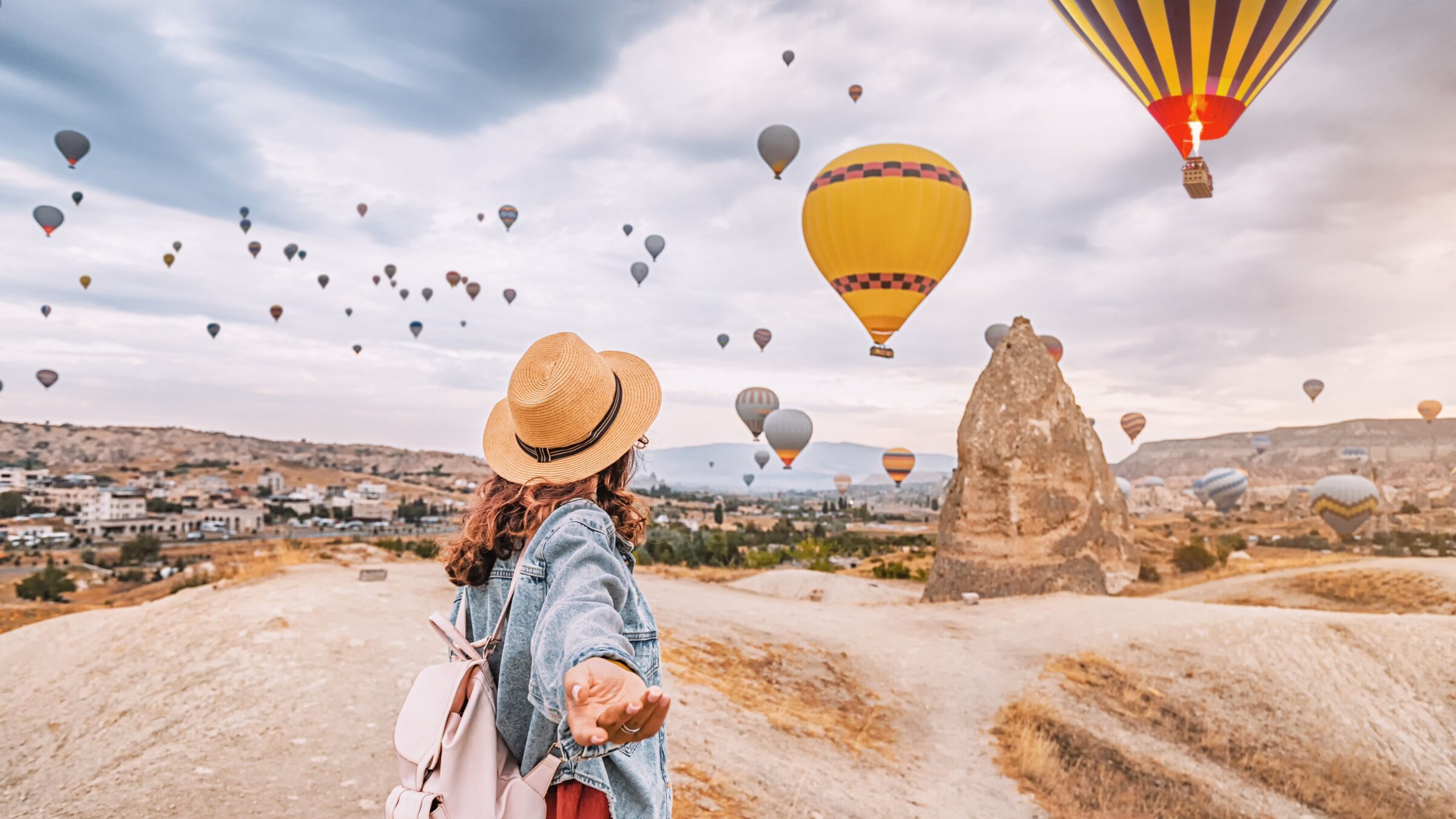 A young woman strikes a confident follow-me pose with hot air balloons soaring in the background.