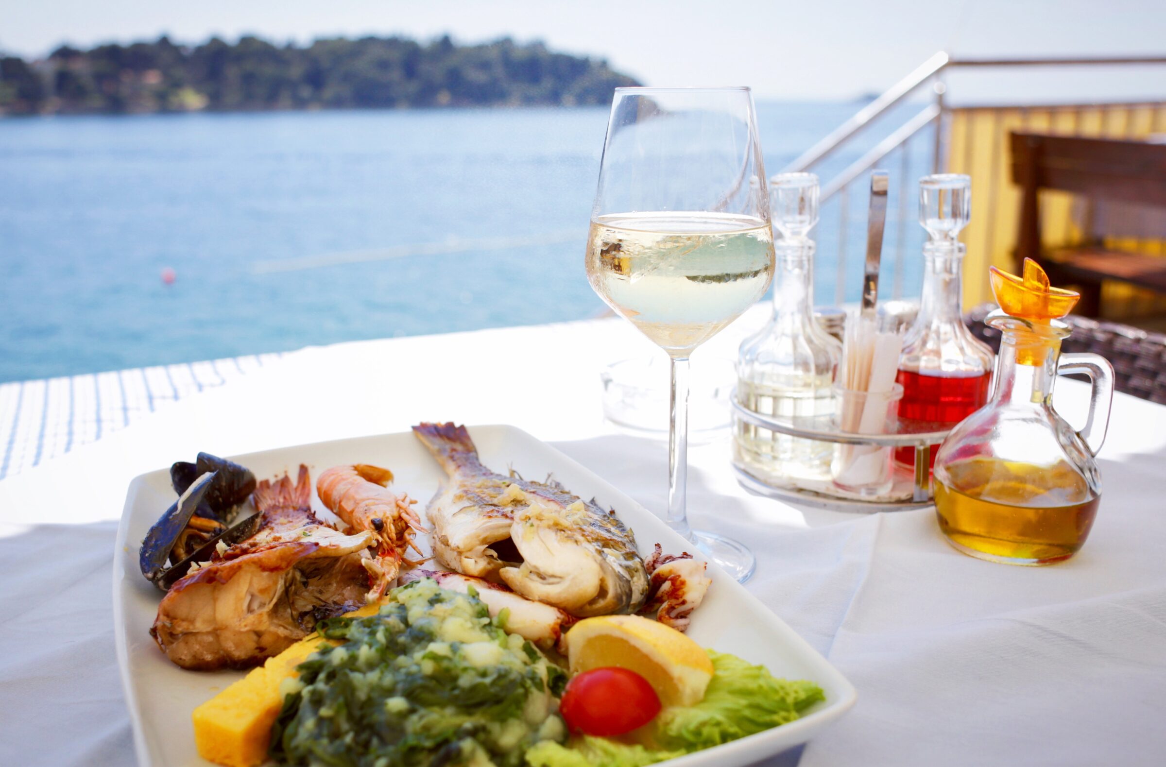 A seafood platter with grilled fish, shrimp, mussels, and vegetables served with a glass of white wine at an outdoor seaside restaurant.