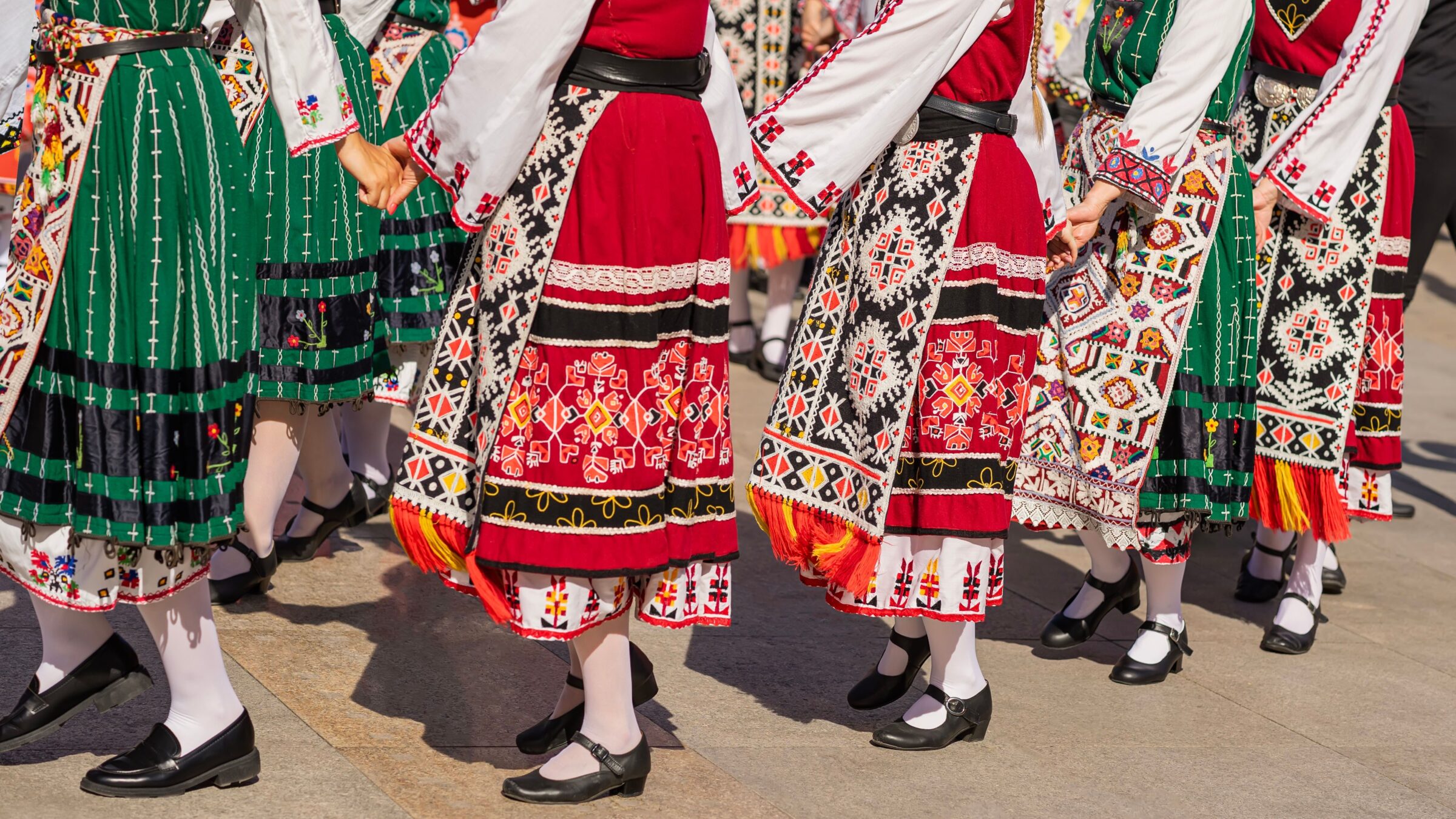 Women in traditional Balkan dresses perform a group folk Bulgarian dance in sunlight. 