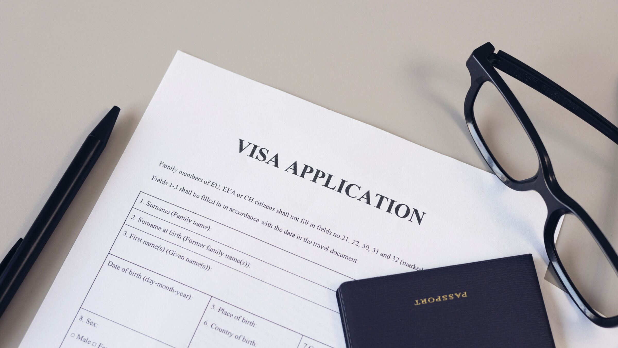 A visa application form, a pen, reading glasses and a passport on a white desk.