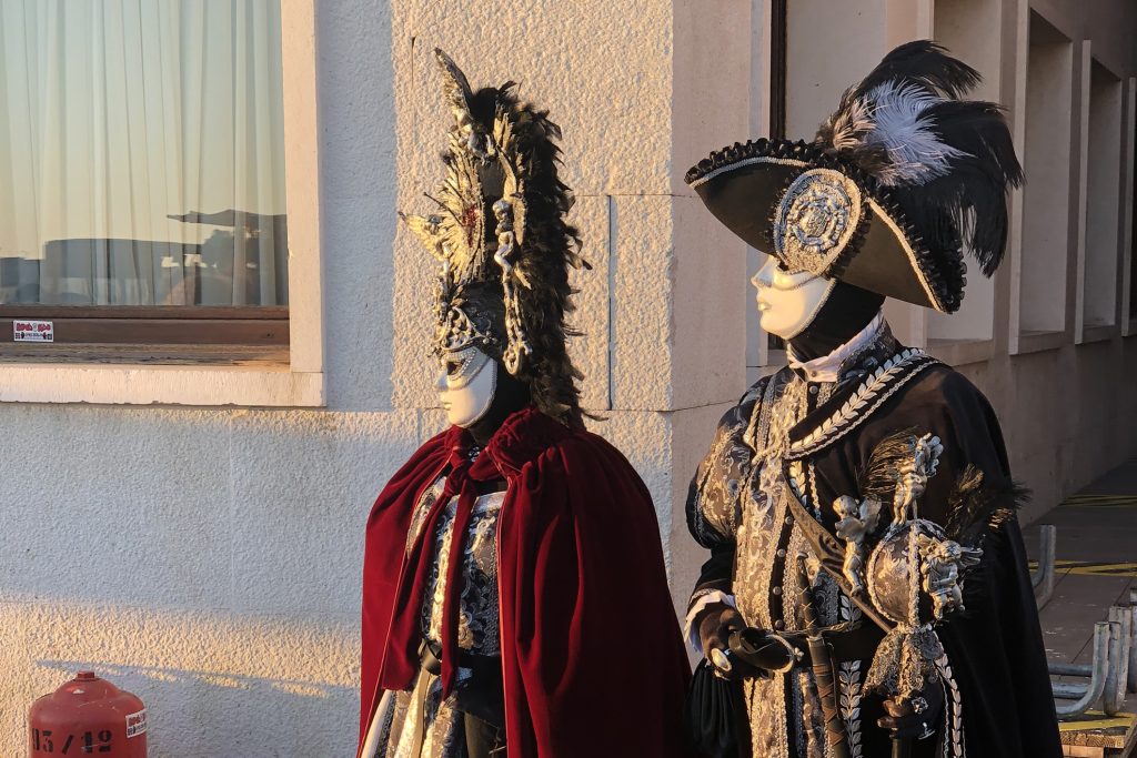 Two people in elaborate Venetian carnival costumes with ornate masks, feathers, and baroque-style embroidery, standing against a light-colored building wall during sunset